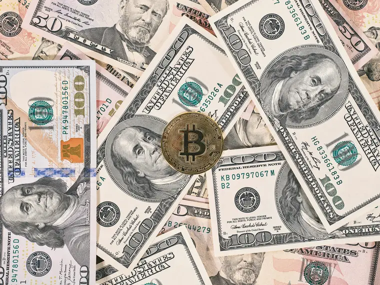 A physical bitcoin token beside banknotes and a smartphone showing a wallet balance